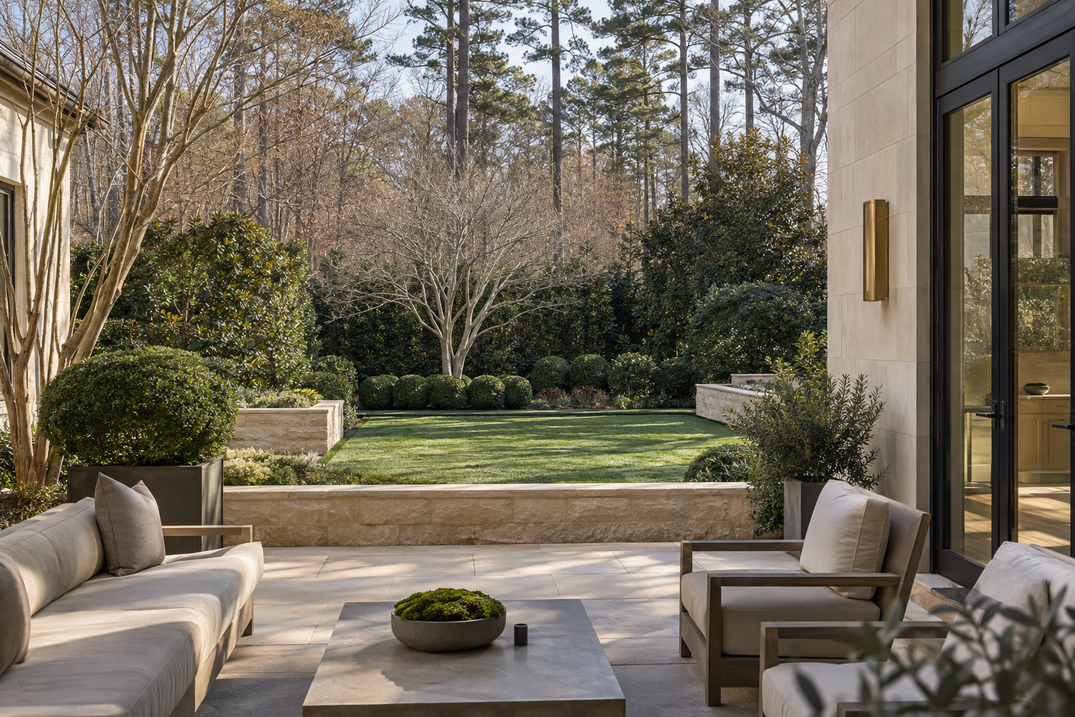 A limestone terrace overlooks a rectangular lawn framed by evergreen screening, a leafless dogwood and mature trees beside a charcoal-framed residence.